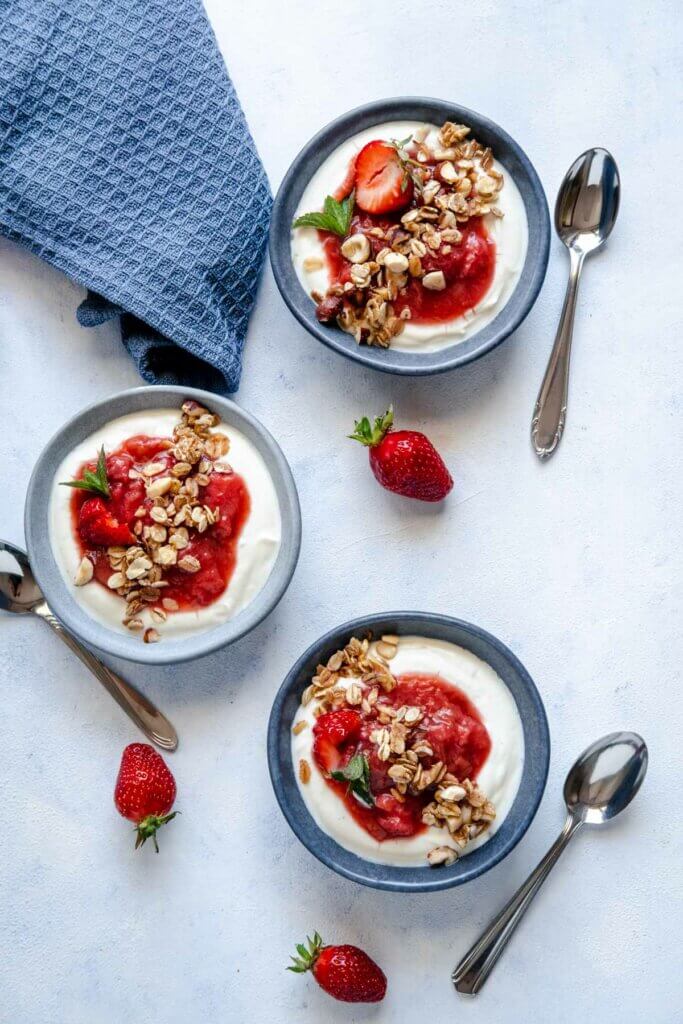 Rhabarber-Erdbeer-Kompott mit Joghurtcreme und Knuspermüsli verteilt auf drei Schüsseln.